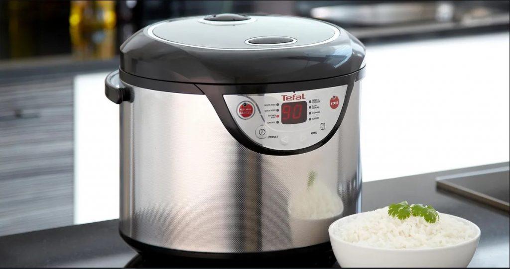 A silver color rice cooker and a bowl of boiled rice at front, a very useful small kitchen appliance. 